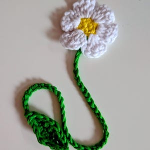 May include: A handmade crochet bookmark featuring a white flower with a yellow center attached to a long, braided green stem. The stem includes a small, textured green leaf detail at the bottom, creating a functional and decorative floral design.