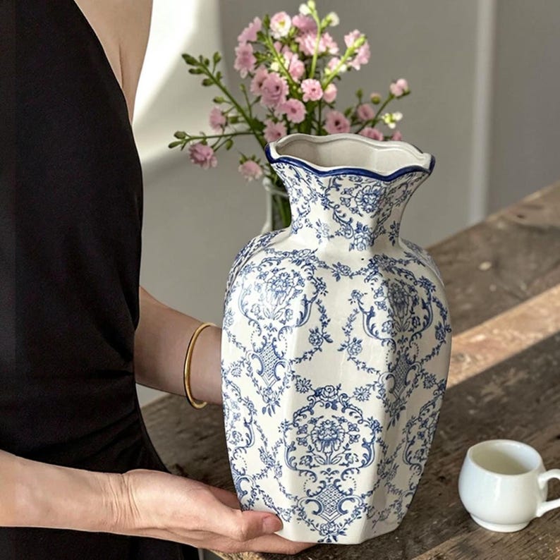 May include: A white ceramic vase featuring a blue floral damask pattern with a scalloped rim. The vase has a hexagonal body shape and a glossy finish. The intricate blue design covers the entire exterior surface of the vessel, contrasting against the white base.