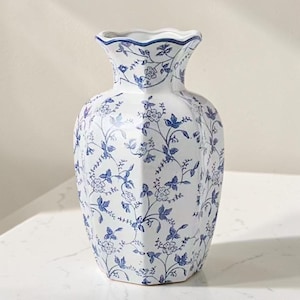 May include: A white ceramic vase with a hexagonal body and a scalloped rim, featuring a repeating blue floral and vine pattern across its entire surface. The design includes delicate blossoms and leaves in a classic blue and white color scheme.