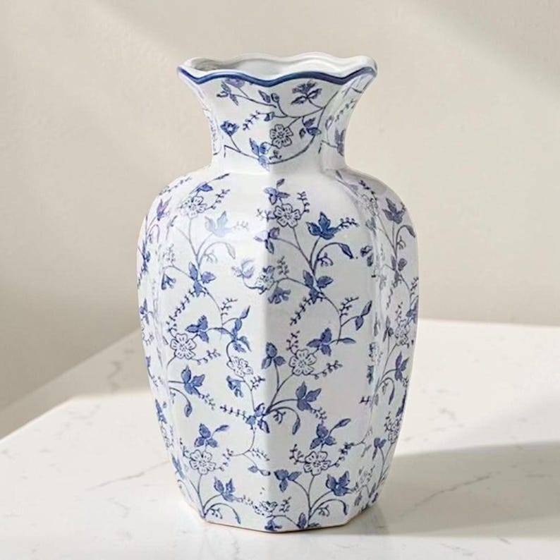 May include: A white ceramic vase with a hexagonal body and a scalloped rim, featuring a repeating blue floral and vine pattern across its entire surface. The design includes delicate blossoms and leaves in a classic blue and white color scheme.