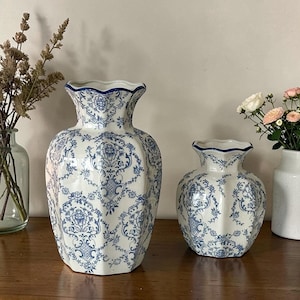May include: Two white ceramic vases featuring a blue floral damask pattern with scalloped rims. The larger vase is tall and bulbous, while the smaller vase is shorter and wider. Both pieces have a glossy finish and a distinct multi-faceted shape.