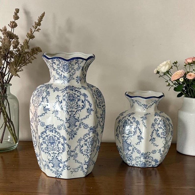 May include: Two white ceramic vases featuring a blue floral damask pattern with scalloped rims. The larger vase is tall and bulbous, while the smaller vase is shorter and wider. Both pieces have a glossy finish and a distinct multi-faceted shape.