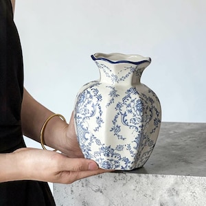 May include: A ceramic vase with a scalloped rim and a hexagonal body, featuring a white base with a detailed blue floral and vine pattern. The design includes intricate scrolling foliage and small blossoms covering the entire exterior surface.
