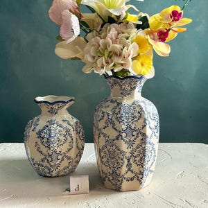 May include: Two cream-colored ceramic vases with a blue floral damask pattern and scalloped rims. The larger vase holds a bouquet of pink, yellow, and white flowers. A small white tag with the letter p is placed between the two vases.
