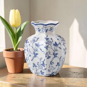 May include: A white ceramic vase with a scalloped rim and a blue floral pattern featuring delicate vines and blossoms. The vase has a hexagonal body shape and is positioned next to a yellow tulip planted in a small terracotta-colored pot.