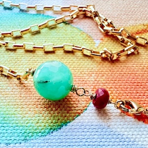 May include: A gold-colored chain necklace with a large, faceted, light green bead and a smaller, round, red bead. The necklace is displayed on a colorful, abstract background.