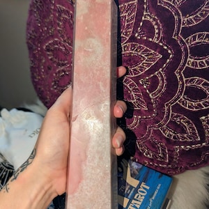 May include: A large, pink quartz obelisk with a pointed top. The stone has a textured surface with variations in color, from light to darker pink. The obelisk is held in a hand, with a tarot card box visible in the background.