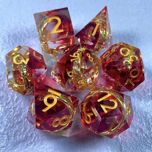 Puede incluir: Juego de siete dados poliédricos. Cada dado es rojo transparente con números y detalles dorados. Los dados están diseñados para juegos de rol de mesa.