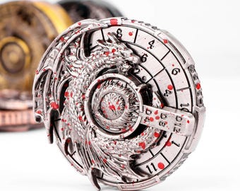 May include: A silver-coloured, round metal object featuring a dragon design and a clock-like face. The dragon is intricately detailed, with red accents. The clock face has numbers and a central dial. Other similar objects are visible in the background.