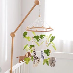 May include: A wooden baby mobile featuring a circular frame with hanging felt jungle animals: a lion, giraffe, zebra, and elephant. Green felt leaves and wooden beads are also suspended, creating a playful nursery decoration.