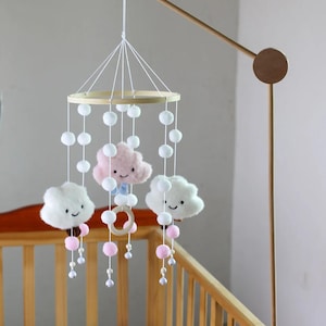 May include: A baby mobile with a wooden frame and hanging plush clouds, pom-poms, and beads. The mobile features three smiling cloud shapes in white and pink, suspended over a wooden crib.