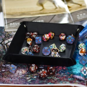 May include: A black leather dice tray containing a collection of multi-sided dice in various colours. The dice are clear, red, blue, and green, with visible numbers. The tray is rectangular and rests on a colourful surface.