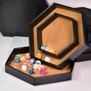 May include: Three black hexagonal dice trays of varying sizes, nested together. The trays have a tan interior and are filled with colorful polyhedral dice. The trays are on a white surface with a black background.