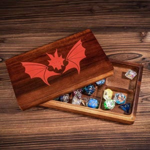 May include: A wooden dice box with a red dragon wing design on the lid. The box is open, revealing various colorful dice inside. The box is made of dark wood and sits on a wooden surface.