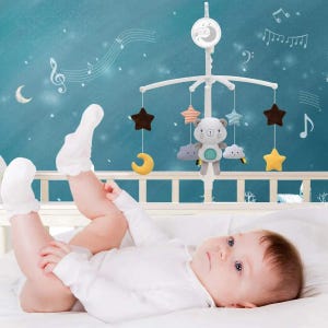 May include: A baby mobile with a white frame, featuring a plush bear, stars, clouds, and crescent moons in various colors. The mobile hangs above a baby in a white onesie, lying on a white bed.