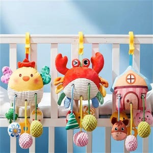 Puede incluir: Tres coloridos juguetes para cuna de bebé colgados de clips de plástico amarillos. Los juguetes incluyen un pollito amarillo, un cangrejo rojo y una casa. Cada juguete tiene elementos colgantes como pompones, campanas y otras formas. Los juguetes están sujetos a una cuna blanca.