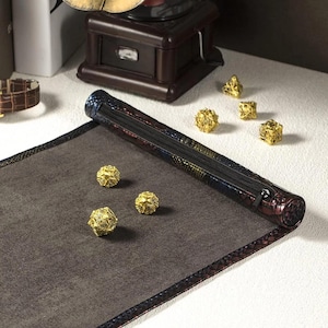 May include: A dark gray dice rolling mat with a patterned border and a rolled-up, zippered case. Several gold-colored, multi-sided dice are scattered on the mat. The case has a dark, textured exterior with blue, red, and yellow accents.