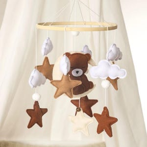Puede incluir: Móvil para bebé con un oso de peluche marrón durmiendo sobre una luna crema. Incluye estrellas marrones y crema, nubes blancas y un aro de madera. Colgando de hilos.
