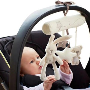 May include: A baby car seat mobile featuring a plush bunny, star, and bear. The toys are cream-colored and suspended from a cream-colored bar with star and stitched designs. The mobile is attached to a black car seat.