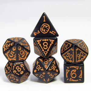 May include: A collection of seven black polyhedral dice featuring gold-coloured engraved numbers and symbols. The dice include a tetrahedron, a cube, a dodecahedron, an icosahedron, and other shapes. The dice are stacked on top of each other.