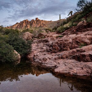 May include: A scenic landscape featuring a rocky desert canyon with a small body of water reflecting the sky. The canyon walls are reddish-brown, and the sky is overcast. Cacti and other desert plants dot the landscape.