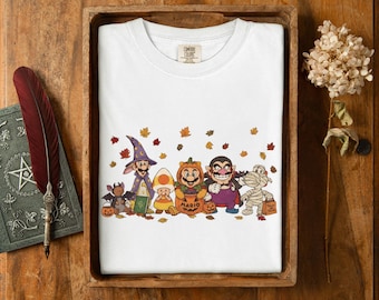Könnte beinhalten: Ein weißes T-Shirt mit einem Grafikdruck verschiedener Videospielcharaktere in Halloween-Kostümen, darunter ein Zauberer, ein Zuckermais, ein Kürbis, ein Vampir und eine Mumie, umgeben von verstreuten Herbstblättern.