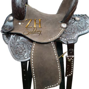 May include: A brown leather saddle with intricate floral and scroll tooling. The saddle features silver accents, white stitching, and the text "ZH Saddlery". The saddle is designed for horseback riding.