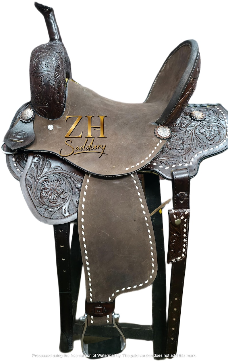 May include: A brown leather saddle with intricate floral and scroll tooling. The saddle features silver accents, white stitching, and the text "ZH Saddlery". The saddle is designed for horseback riding.