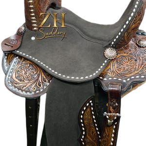 May include: A Western-style saddle with a dark gray seat and intricate floral tooling on the brown leather. The saddle features white stitching, silver accents, and the "ZH Saddlery" logo.