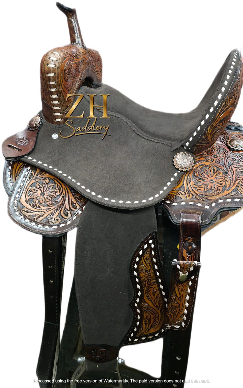 May include: A Western-style saddle with a dark gray seat and intricate floral tooling on the brown leather. The saddle features white stitching, silver accents, and the "ZH Saddlery" logo.