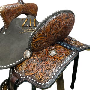 May include: A brown leather western saddle with intricate floral tooling. The saddle features a dark gray seat, silver conchos, and white stitching. The saddle is designed for horseback riding and is a traditional style.