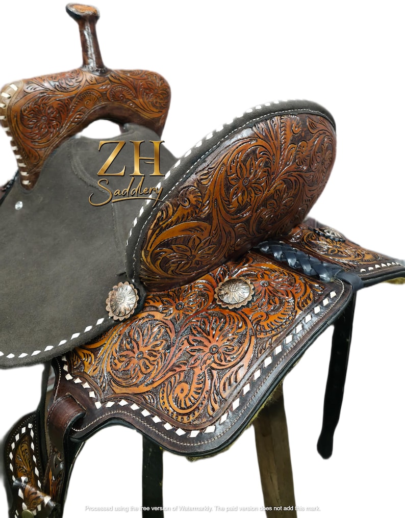 May include: A brown leather western saddle with intricate floral tooling. The saddle features a dark gray seat, silver conchos, and white stitching. The saddle is designed for horseback riding and is a traditional style.