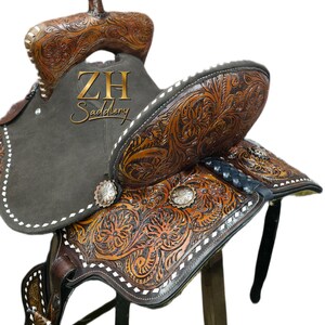 May include: A brown leather saddle with intricate floral carvings and white stitching. The saddle features a dark gray suede seat and silver-toned conchos. The text "ZH Saddlery" is visible on the saddle.