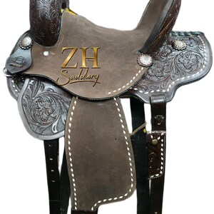 May include: A brown leather Western saddle with intricate floral tooling and silver accents. The saddle features a high cantle, a horn, and stirrups. The text "ZH Saddlery" is visible on the saddle. The saddle is designed for horseback riding.