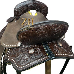May include: A brown leather western saddle with intricate floral tooling and white stitching. The saddle features a high cantle, a horn, and stirrups. The text "ZH Saddlery" is visible on the saddle.