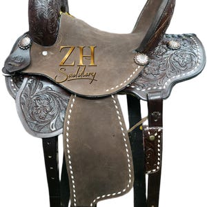 May include: A brown leather Western saddle with intricate floral tooling and silver accents. The saddle features a high cantle, a horn, and stirrups. The text "ZH Saddlery" is visible on the saddle. The saddle is designed for horseback riding.