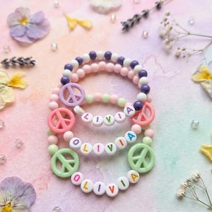 Può includere: Tre braccialetti elastici di perline con perline bianche rotonde che compongono il nome OLIVIA in lettere colorate. Ogni braccialetto include due ciondoli a forma di segno di pace in tonalità pastello viola, rosa e verde chiaro, con perline.