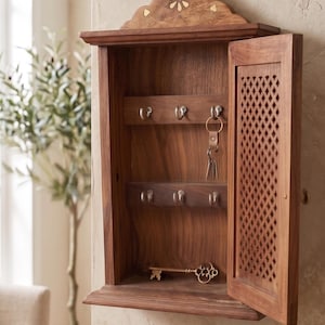 May include: A wooden key cabinet with a decorative floral design on top and a lattice-patterned door. The cabinet is open, revealing key hooks and a decorative key. The wood has a warm, natural finish.