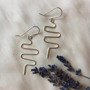 May include: A pair of silver-colored, abstract wave-shaped earrings with hook closures. The earrings are displayed on a white fabric surface, with a sprig of dried lavender in the lower right corner. The earrings have a minimalist, modern design.