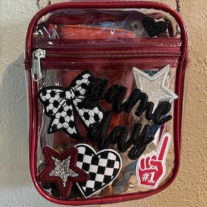 May include: Clear, rectangular crossbody bag with a red trim and zip. The bag is decorated with patches including a soccer ball bow, a glittery star, a checkered heart, and the words "Game Day".
