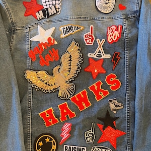 May include: Light blue denim jacket adorned with various patches. Patches include a gold sequined eagle, red stars, the word "HAWKS", and phrases like "Game Day" and "Raising Legends".