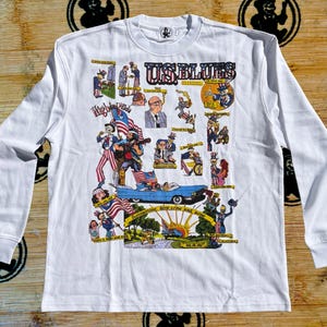 Puede incluir: Una camiseta blanca de manga larga con un estampado gráfico multicolor titulado U.S. BLUES. El diseño incluye varias ilustraciones de dibujos animados del Tío Sam, un coche descapotable azul, banderas estadounidenses y texto como Summertime.