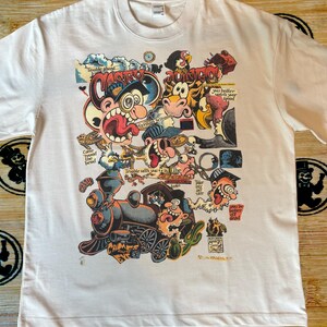 Puede incluir: Camiseta de color crema con un gráfico de dibujos animados colorido. El diseño incluye un tren, varios personajes y el texto "Trouble Ahead" y "Casey Jones". Las frases incluyen "Come round the bend" y "You better watch your speed".