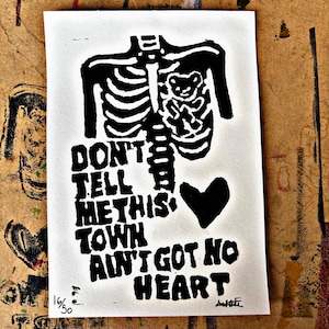 May include: Black and white print featuring a skeleton with a teddy bear in its ribcage and the text "DON'T TELL ME THIS TOWN AIN'T GOT NO HEART." A black heart is next to the text. The print is numbered 16/50.