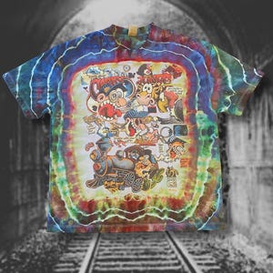 Puede incluir: Una camiseta de manga corta con estampado tie-dye que presenta un gráfico colorido y caótico de personajes de dibujos animados, una locomotora de vapor y varios elementos de texto como "Casey Jones" y "You better watch your speed" en un patrón multicolor.