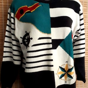 May include: A white, black, and teal striped sweater with a nautical theme. The sweater features a red and gold flag, a ship's wheel, and a compass.
