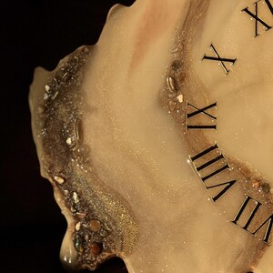 May include: A unique, handmade clock with a resin finish. The clock face features a creamy white and gold design with embedded stones and glitter. Roman numerals mark the hours. The clock has an irregular, organic shape.