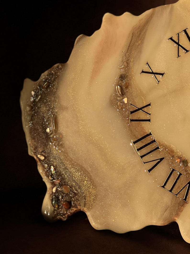 May include: A unique, handmade clock with a resin finish. The clock face features a creamy white and gold design with embedded stones and glitter. Roman numerals mark the hours. The clock has an irregular, organic shape.