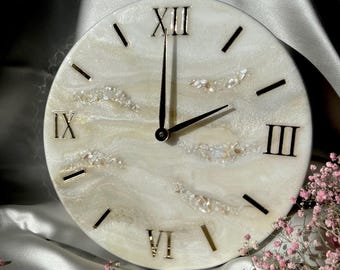 May include: Round wall clock with a white and gold marbled design. The clock face features Roman numerals and gold-colored hands. Decorative accents include small, iridescent pieces. The clock is set against a satin background.