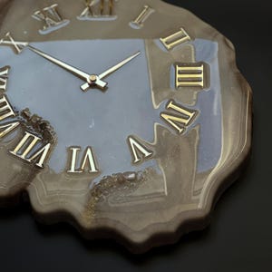 May include: A unique wall clock with a natural, organic shape. The clock face is a muted gray with gold Roman numerals and hands. The clock's edge has a marbled, brown and gold resin finish, creating a striking contrast.